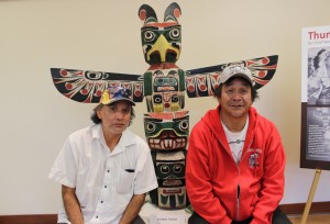 Two men sitting in front of a colourful thunderbird totem pole.