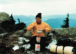 Man siting outdoors eating scrambled eggs from a pan with mountains in the background.