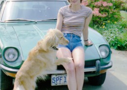 Woman leaning against the hood of her car with a dog jumping up onto her.