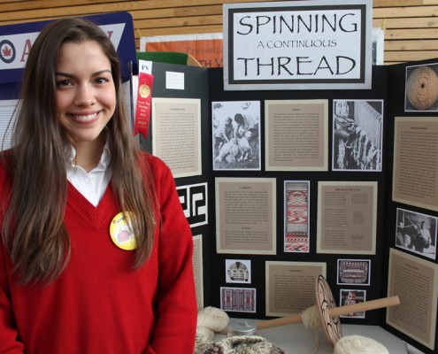 Young girl posing with her Heritage Fair project, with weaving artifacts on the table beside her.
