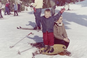 Woman kneeling next to a boy who is standing on a “flying saucer” sled, his arms up in the air.