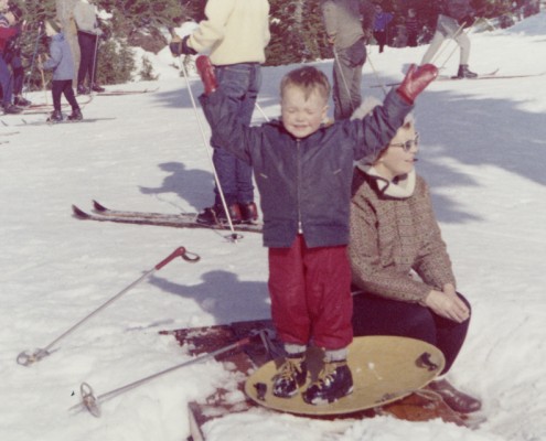 Woman kneeling next to a boy who is standing on a “flying saucer” sled, his arms up in the air.