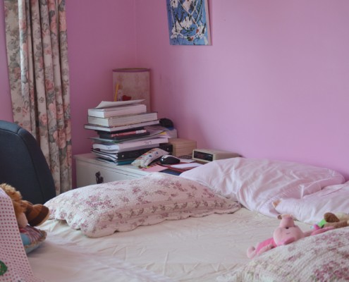 Corner of a bedroom, showing unmade bed, pillows, pink walls, and a stack of books.