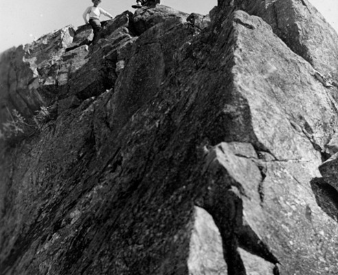 View from below of two men sitting on the rocky ridge of a mountain.