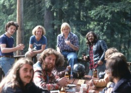 Several men sitting around a picnic table on a deck, trees in the background.