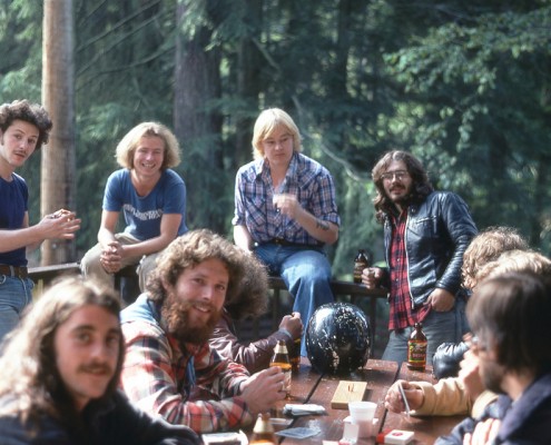Several men sitting around a picnic table on a deck, trees in the background.