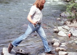 Man with fishing rod stepping from one rock to another in a stream. 5tags: photography, North Vancouver, Lynn Valley, Bruce Flanagan, softball, archives