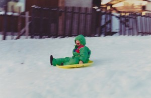 Young boy wearing a snowsuit, sitting on a sled, a smile is on his face.