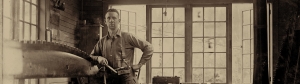 Man in his windowed workshop stops sharpening a tree saw to pose for a picture. His dress of button down work shirt and suspender along with his surrounding suggests early to mid-1900’s