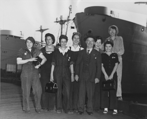 First women to be employed at Burrard Dry Dock in sheet metal shop during World War 2. Front row, L.to R: ?, Eleanor Morfitt, Robert Logan, ?. Back row: ?, ?, ?, Claudia Eckstein. Victory ships in background. Photo: NVMA 12384