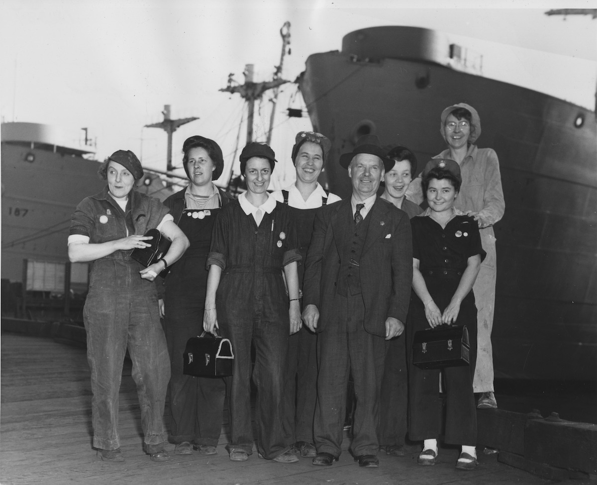 First women to be employed at Burrard Dry Dock in sheet metal shop during World War II. Front row, L.to R: ?, Eleanor Morfitt, Robert Logan, ?. Back row: ?, ?, ?, Claudia Eckstein. Victory ships in background. Photo: NVMA 12384